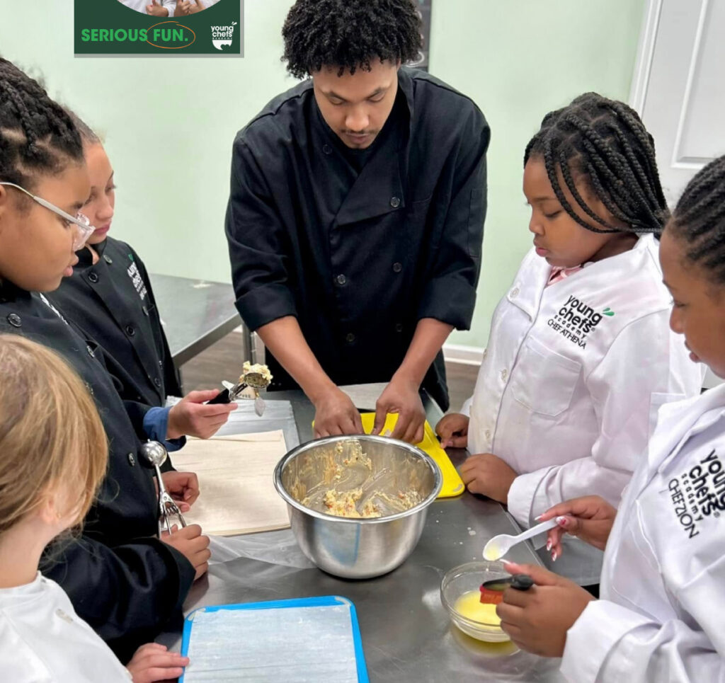 A Young Chefs Academy instructor guiding kids in chef coats during a cooking class